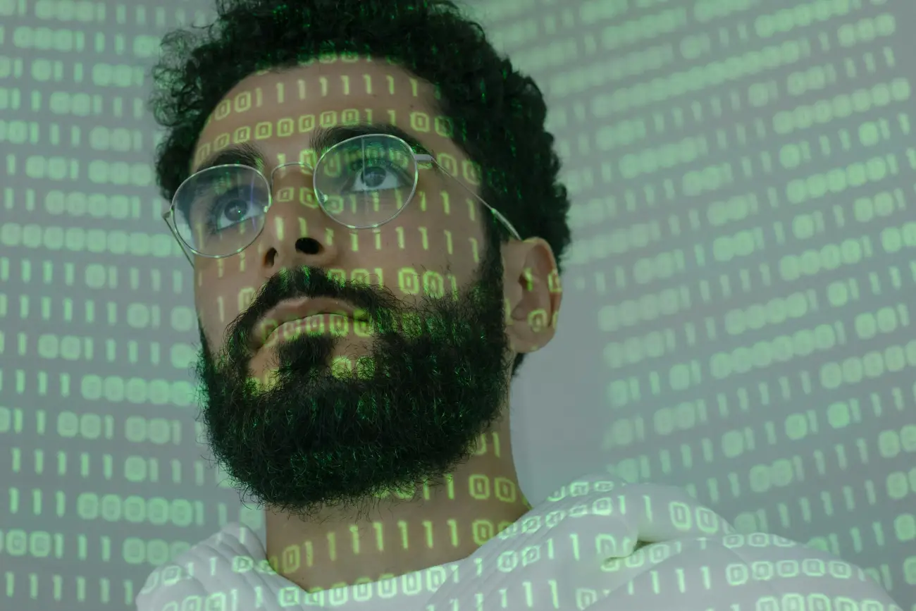 A portrait of a man with glasses and a beard, with green binary code (0s and 1s) projected across his face and background.