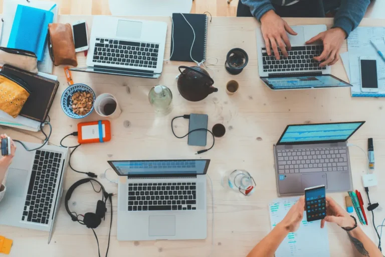 An overhead view of a wooden desk filled with several laptops, smartphones, notebooks, and people working together.