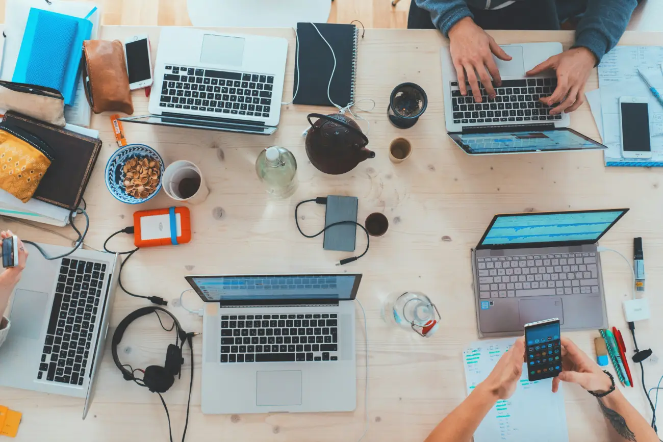An overhead view of a wooden desk filled with several laptops, smartphones, notebooks, and people working together.