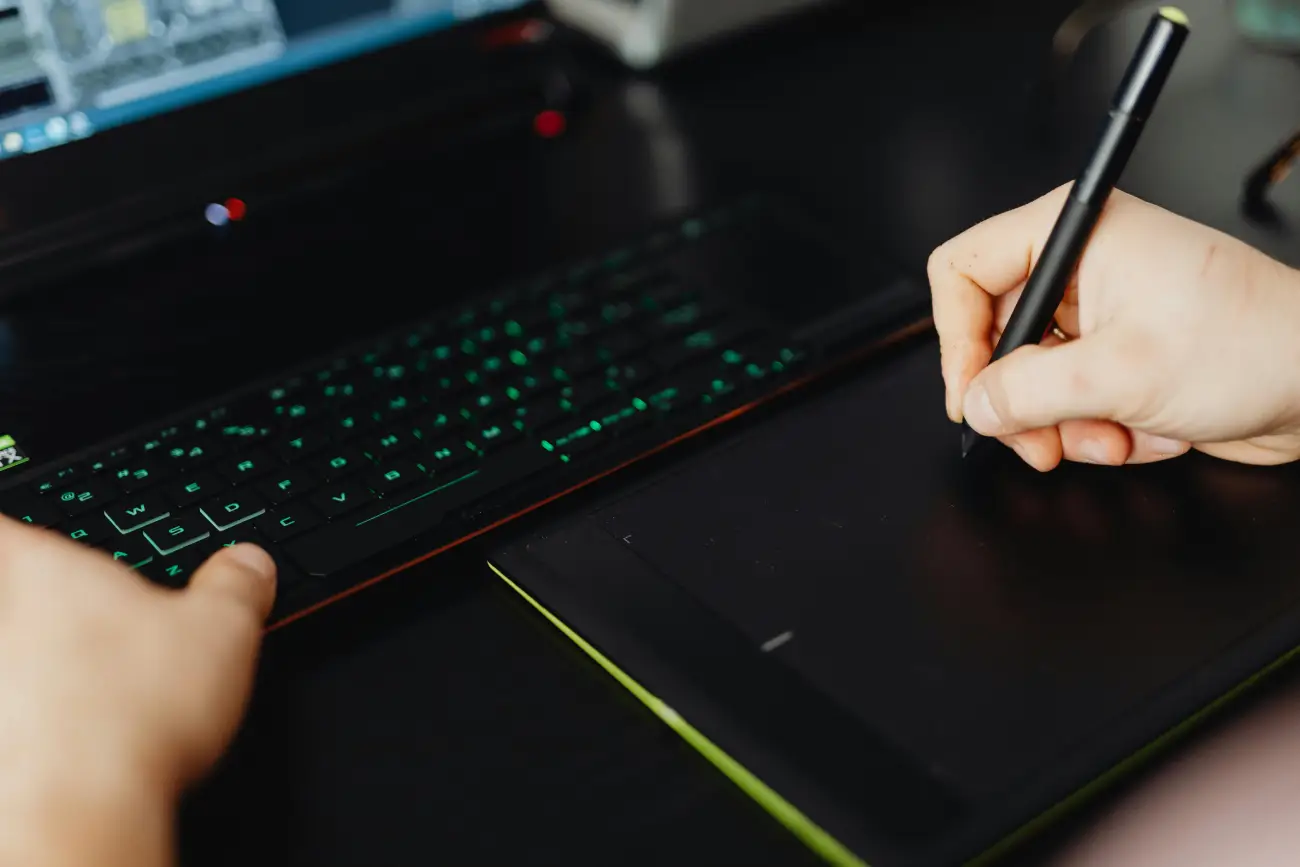 A high-angle shot of a person using a black digital drawing tablet and a stylus next to a backlit laptop keyboard.